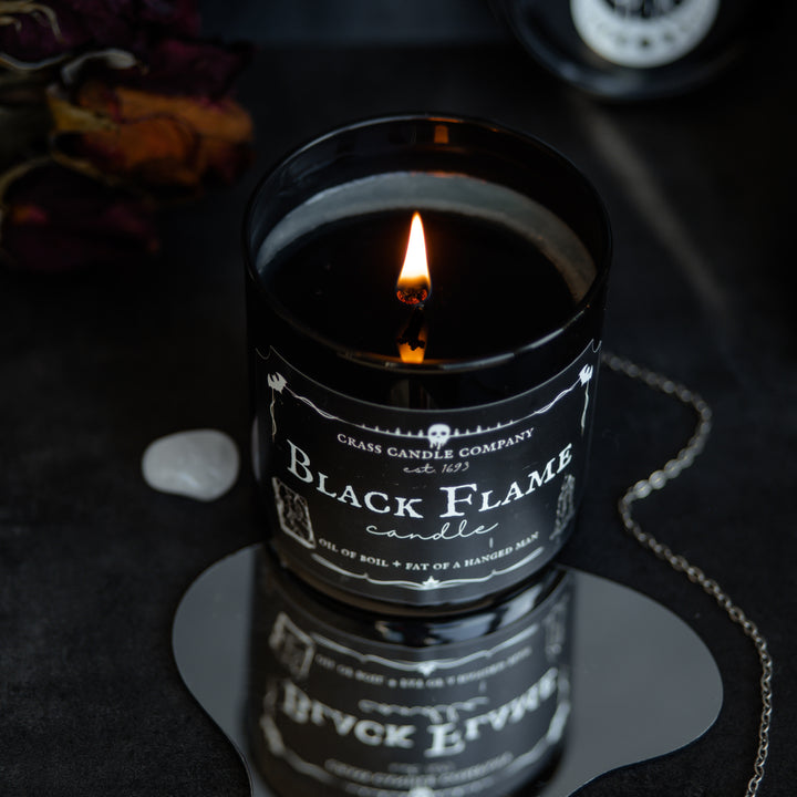 A shot of the Black Flame Candle jar from above. The black wax is melted and the flame glows bright. The candle sits on a mirror that shows a different angle. Next to it is a crystal and a pendulum.