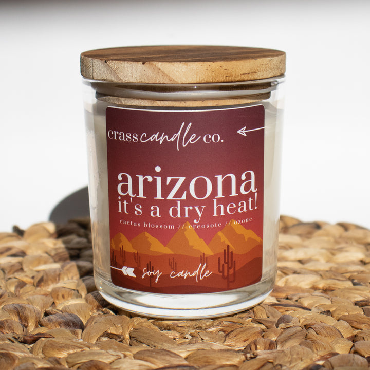 A clear jar filled with white soy wax, topped with a smoky wood lid. The jar features a label that reads "Arizona, It's a Dry Heat" with a sunset backdrop, mountains, and cacti in the foreground. At the top of the label displays the Crass Candle Co. logo.