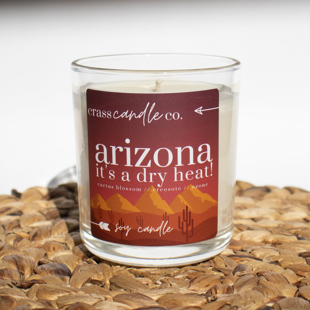 A clear jar filled with white soy wax, topped without the smoky wood lid. The jar features a label that reads "Arizona, It's a Dry Heat" with a sunset backdrop, mountains, and cacti in the foreground. At the top of the label displays the Crass Candle Co. logo.