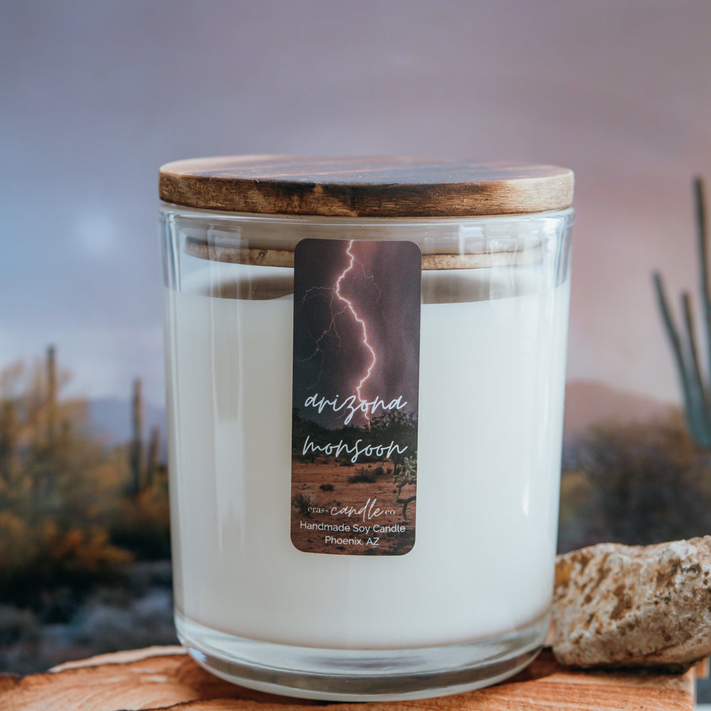 A clear jar filled with white soy wax, topped with a smoky wood lid. The jar features a small vertical, rectangle label that reads "Arizona Monsoon" with a desert backdrop and lightening in the foreground. The bottom of the label displays the Crass Candle Co. logo.