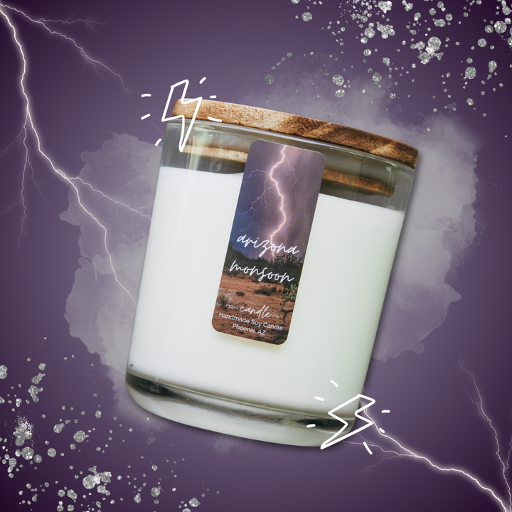 A clear jar filled with white soy wax, topped with a smoky wood lid. The jar features a small vertical, rectangle label that reads "Arizona Monsoon" with a desert backdrop and lightning in the foreground. The bottom of the label displays the Crass Candle Co. logo. The candle is on a purple background with lightning graphics.