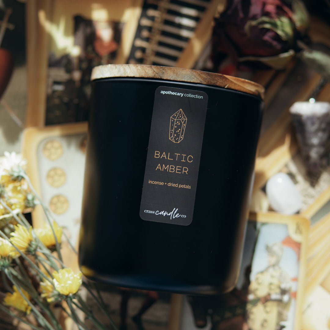 A black jar sitting on top of tarot carts, topped with a smoky wood lid. The jar features a vertically placed rectangle label that reads "Baltic Amber" in copper lettering with a gemstone graphic. The label displays the Crass Candle Co. logo, the words "Apothecary Collection" and the scent notes: amber and musk.