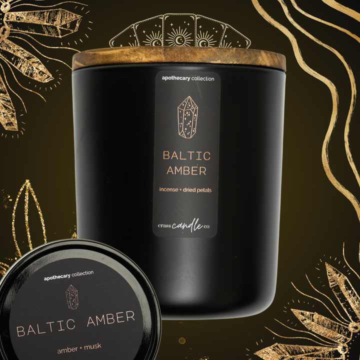 A black jar sitting on top of a green and gold graphic with tarot cards and swirls, topped with a smoky wood lid, as well as a black round tin lid. The jar features a vertically placed rectangle label that reads "Baltic Amber" in copper lettering with a gemstone graphic. The label displays the Crass Candle Co. logo, the words "Apothecary Collection" and the scent notes: amber and musk.