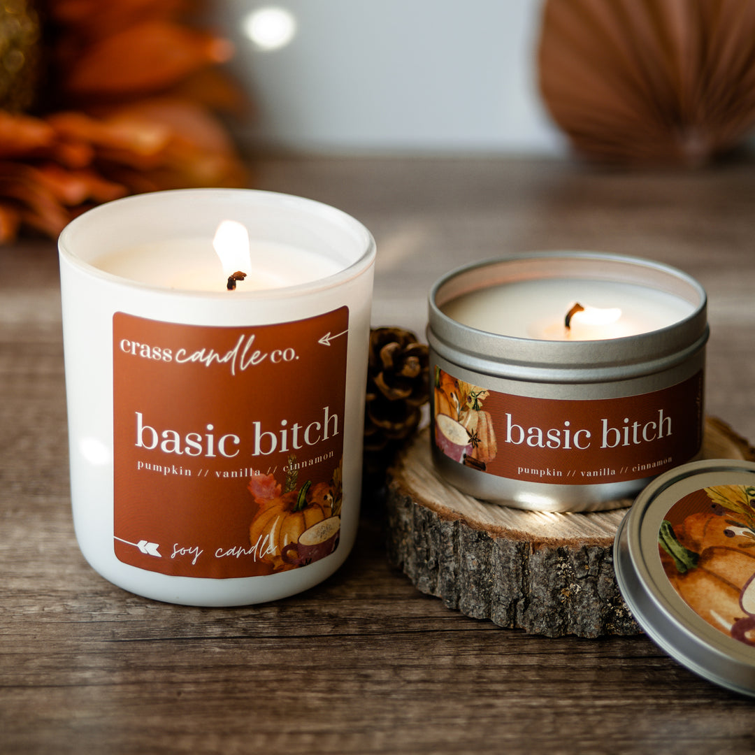 A large white jar with a deep orange square label that says "Basic Bitch" with watercolor pumpkins and a latte in the corner. Next to the jar is a tin sitting on a piece small piece of wood with the lid leaning against it. Both candles are lit. 