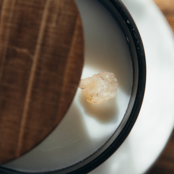 An up close shot of the top of the candle with white wax and a smoky wood lid ajar. The candle has a small crystal on top of it.