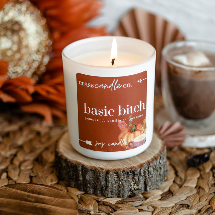 A lit candle in a white jar with a square orange label that says "Basic Bitch" in white lettering. The jar sits on a small piece of wood surrounded by cozy fall items like a mug of hot chocolate and orange flowers. Is there Baileys in the mug? Probably.