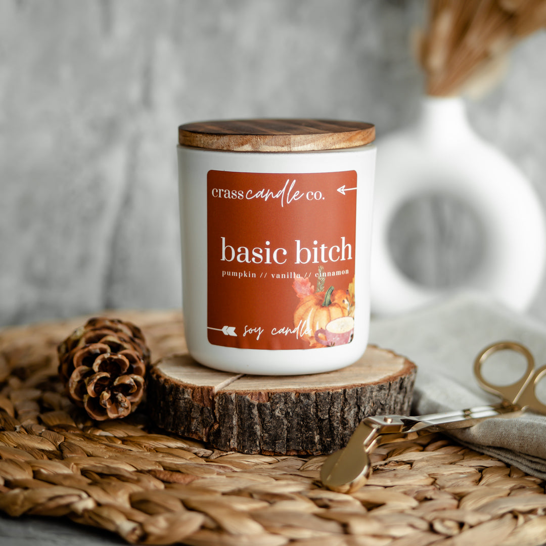 A white jar with the lid on with a square orange label that says "Basic Bitch" in white. Next to the jar is a set of gold wick trimmers and a pinecone.