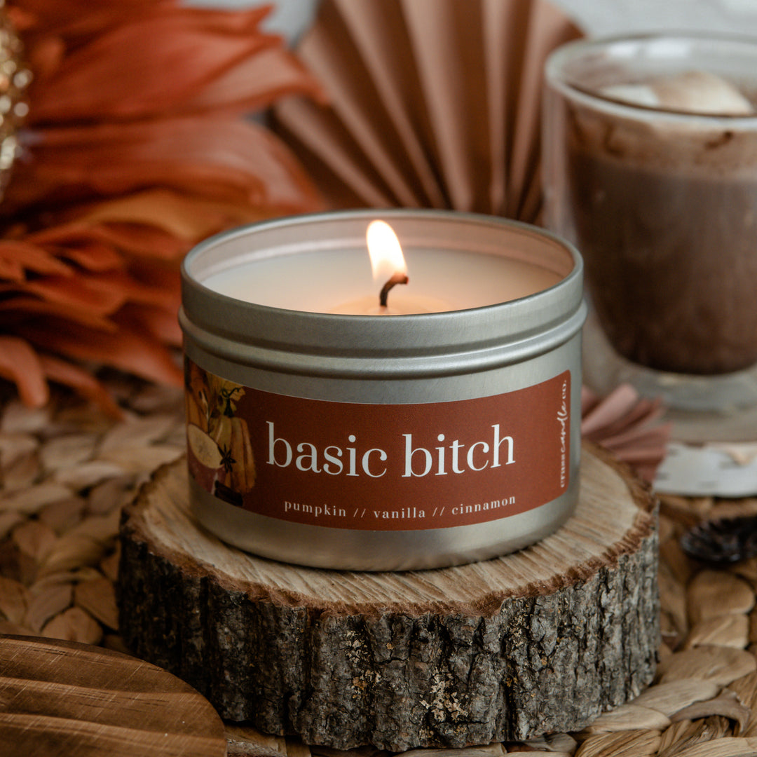 A lit candle in a silver tin with a rectangle orange label that says "Basic Bitch". The candle sits on a small piece of wood next to a cup of hot chocolate. Undetermined if the mug has any booze in it.