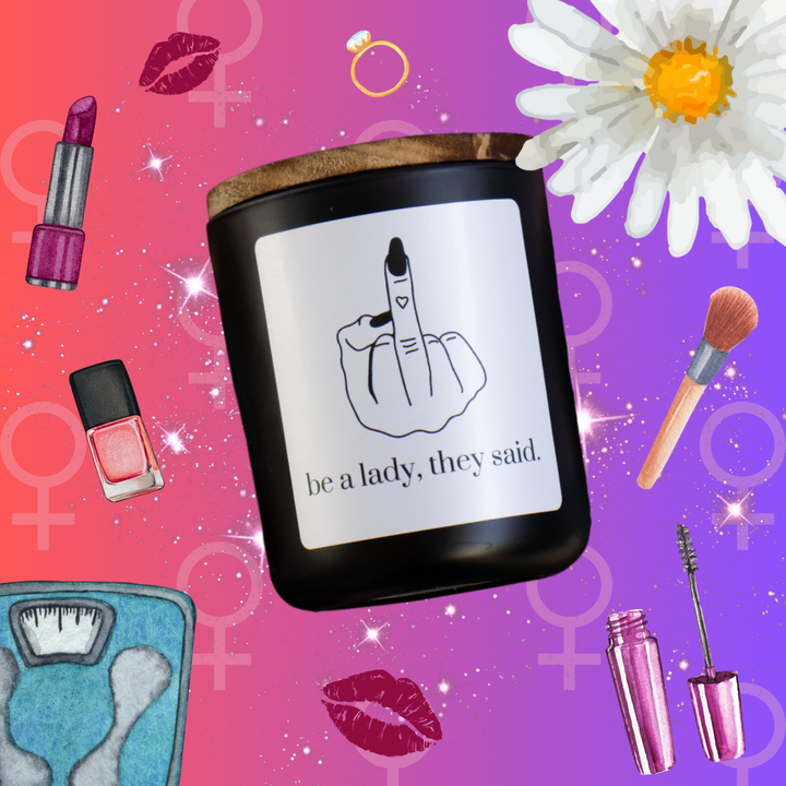 A black jar filled topped with a smoky wood lid. The jar features a white label that has a black outline of a woman's middle finger. The middle finger has black fingernails and a small heart tattoo. Underneath the hand are the words "Be a Lady, They Said" in black. Surrounding the candle are feminine items such as lipstick, flowers, makeup, a scale and nail polish.