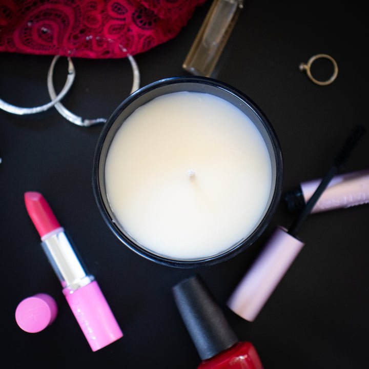 A shot of the candle from the top showing a black jar and white wax with feminine items strewn about next to it such as lipstick and nail polish.