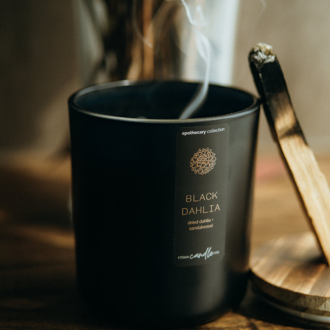 A black jar candle has smoke coming out of it as if its just been blown out. A piece of palo santo leans up against the candle. The black jar has a rectangle label placed vertically which says "Black Dahlia" in copper lettering.