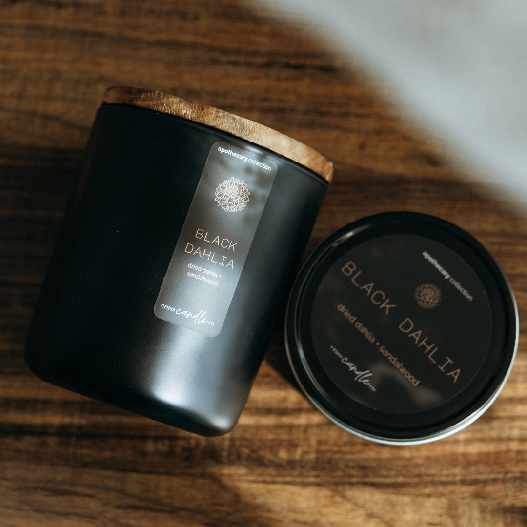 A black jar with a black rectangle label placed vertically on the jar which has copper lettering that says "Black Dahlia", copper dahlia graphic and scent notes that say "dried dahlia and sandalwood".  The candle has its lid on and sits next to a black round tin candle with a similar label, only round. Both candles sit on a wood table.