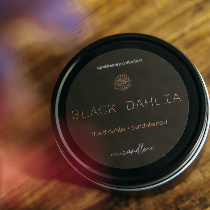 A black, round tin with a round black label. The label says "Black Dahlia" in copper lettering with scent notes "Dried dahlia and sandalwood". At the bottom of the label is a white Crass Candle Co. logo. At the top it says Apothecary Collection.