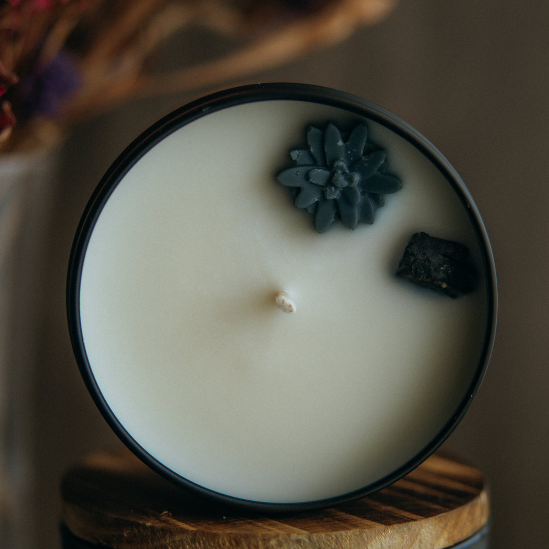 A black tin sits on its side with the lid off. You can see the white soy wax and wick, with a small black wax flower and tourmaline stone. In the background is flowers.