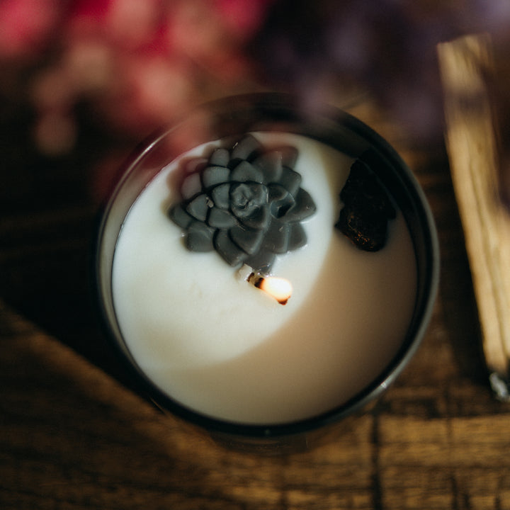 View from the top of the candle which shows white wax inside a black jar. The white wax holds a black wax flower on top, with a small piece of black tourmaline next to it. The candle is lit and the wax is starting to melt. There's a stick of palo santo nearby.