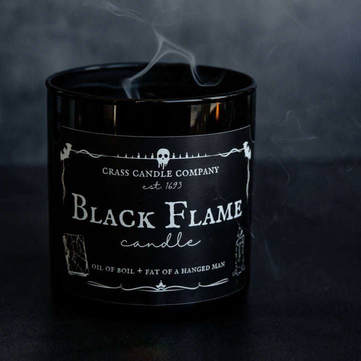 A Black Flame Candle jar has recently been blown out and the smoke is creeping out from the jar. The candle sits against a black background.