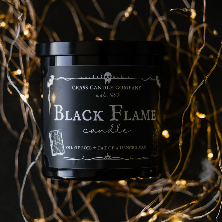 A Black Flame Candle jar sits on a black background with twinkle lights. The lid is on and a light comes from the left side. The label is black with white lettering and spooky graphics.