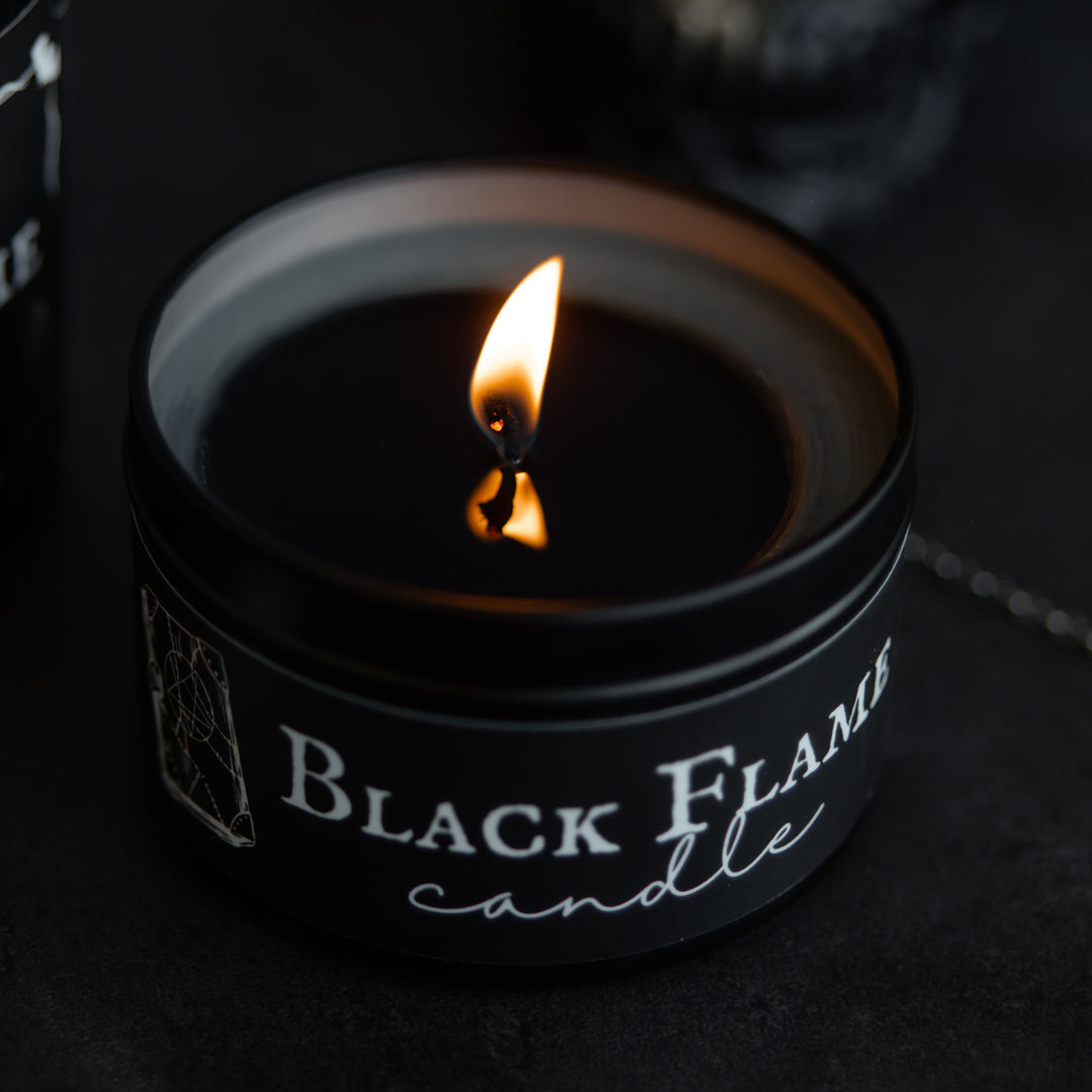 The wick of the Black Flame Candle tin is lit and the black melted wax is starting to reach the perimeter of the tin. The candle sits against a black background. The flame reflects off the black melted wax.