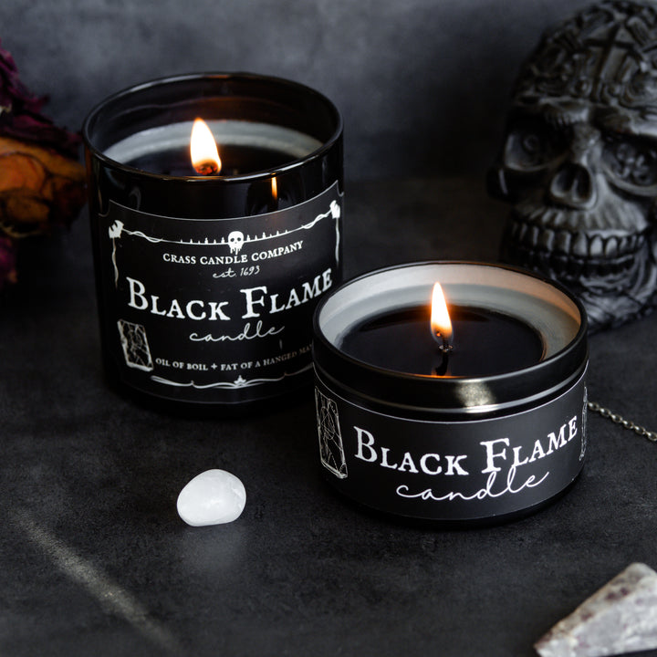 Two candles sitting on a black backdrop next to a black skull figure, whie stone, pendulum and dried rose. One candle is a black jar with a rectangle label that reads "Black Flame Candle" in white lettering. The perimeter of the label has spooky white graphics including a Spell Book, bats, and a candle. Next to the jar is a black tin with a similar label.