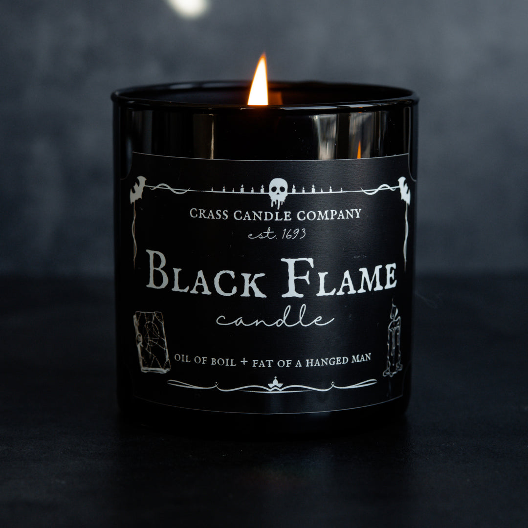 A single Black Flame Candle against a black background. The candle is lit. The front shows a black rectangle label with white lettering and spooky graphics.