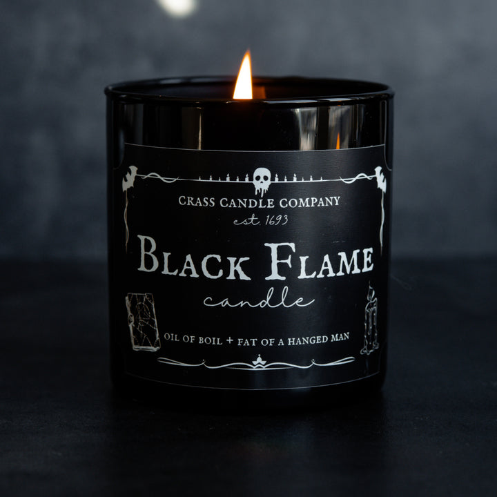 A single Black Flame Candle against a black background. The candle is lit. The front shows a black rectangle label with white lettering and spooky graphics.