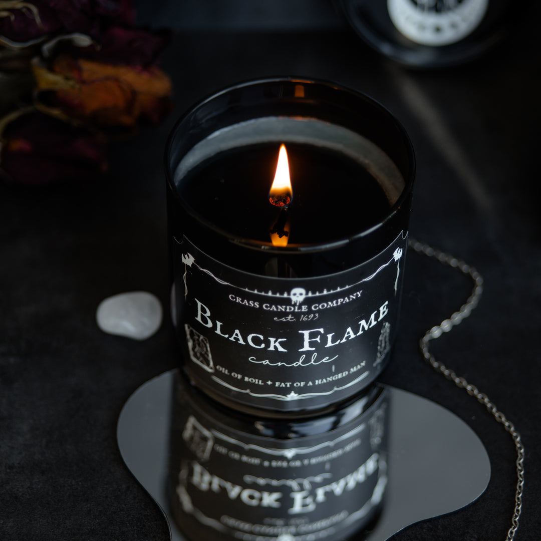 A shot of the Black Flame Candle jar from above. The black wax is melted and the flame glows bright. The candle sits on a mirror that shows a different angle. Next to it is a crystal and a pendulum. 