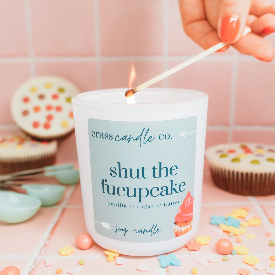Candle labeled 'crass candle co. shut the fucupcake' being lit with colorful sprinkles and cupcakes in the background.