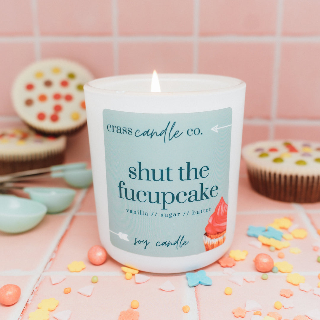 Candle with 'shut the fucupcake' text on a pink tiled background with cupcakes and sprinkles.
