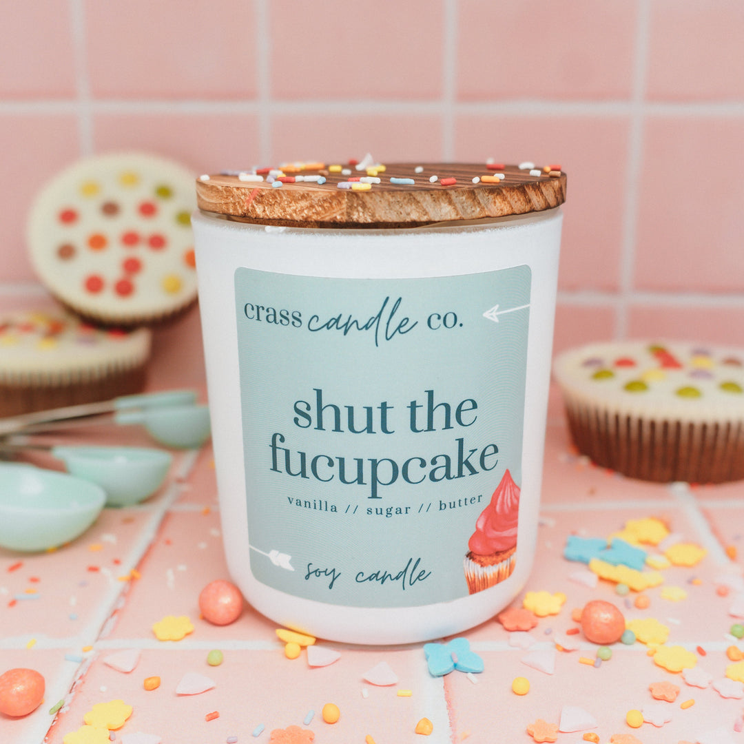 Candle with a humorous label on a surface with colorful confetti and cupcakes.