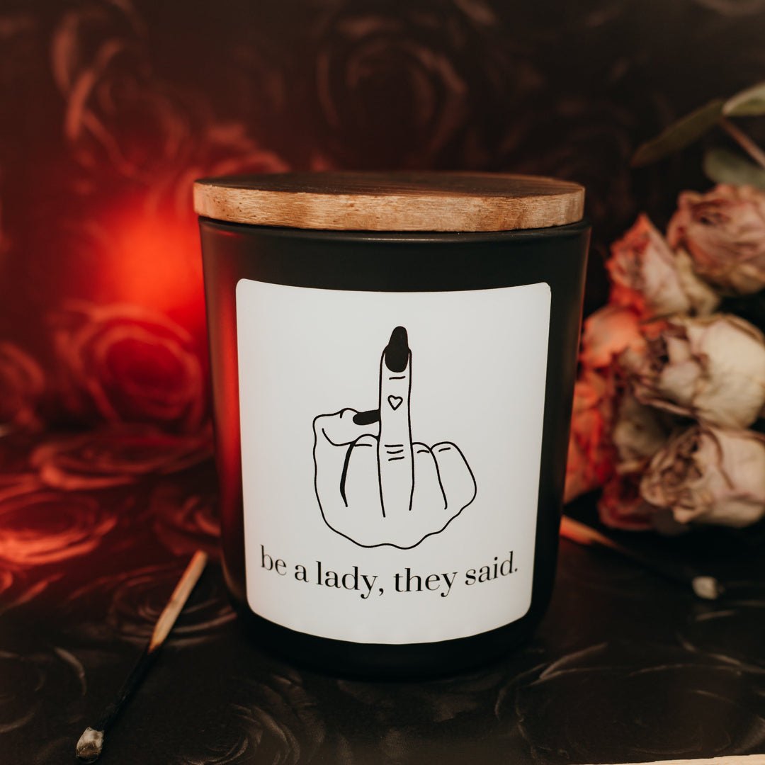 Candle with a wooden lid and a humorous label surrounded by flowers and leaves.