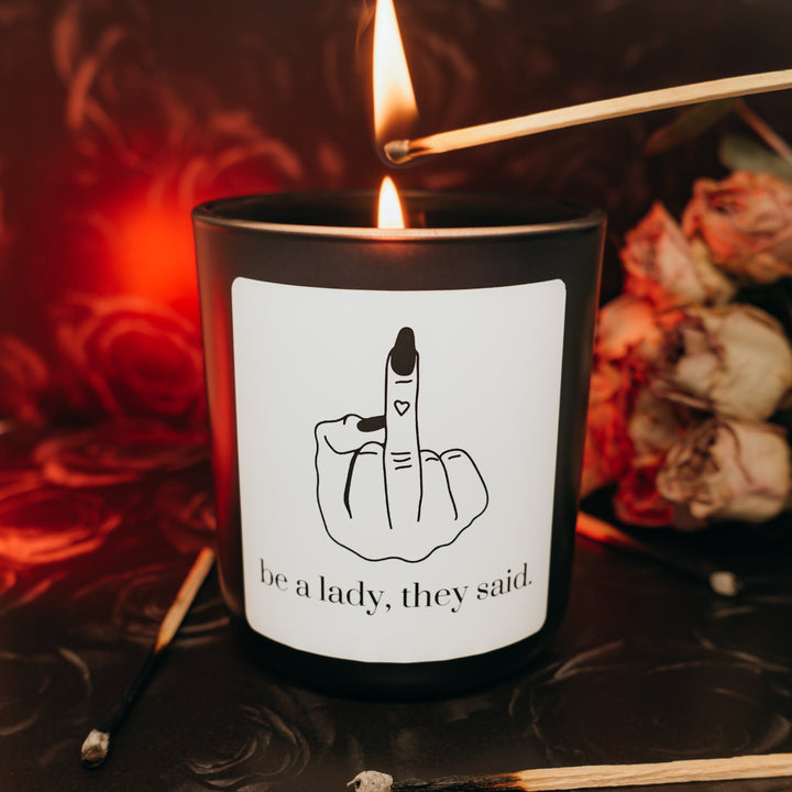 Candle with a label featuring a hand gesture and text, surrounded by dried flowers and matches on a dark background.